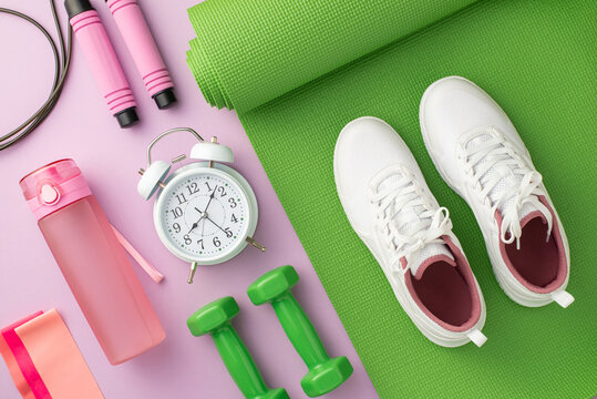 Sports concept. Top view photo of white sneakers over green exercise mat dumbbells alarm clock skipping rope pink bottle and resistance bands on isolated pastel lilac background