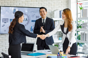 Two happy millennial professional successful businesswomen in formal suit standing shaking hands greeting together when business deal agreement done while businessman entrepreneur smiling clapping