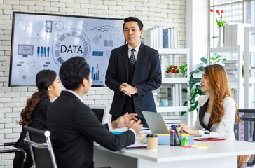 Asian millennial professional successful businessman in black formal suit standing presenting showing company data on big monitor screen with male and female colleagues in meeting room workstation