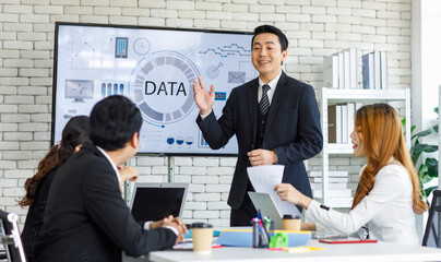 Asian millennial professional successful businessman in black formal suit standing presenting showing company data on big monitor screen with male and female colleagues in meeting room workstation