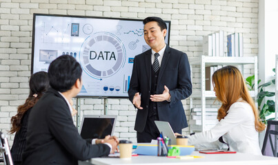 Asian millennial professional successful businessman in black formal suit standing presenting showing company data on big monitor screen with male and female colleagues in meeting room workstation