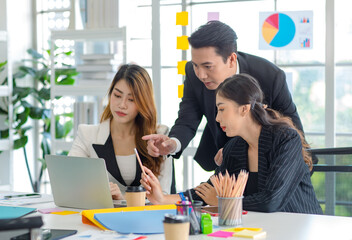 Asian millennial professional successful male businessman mentor in black formal suit standing helping two female employee sitting working with laptop computer at workstation in company meeting room