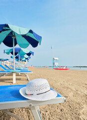 Obraz premium White straw hat on a beach bed with colorful umbrellas on a sunny summer day. In the background a lifeboat alongside a lifeguard tower on the shore of the Adriatic Sea, Italy.