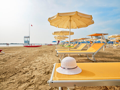 White straw hat on a beach bed with colorful umbrellas on a sunny summer day. In the background a lifeboat alongside a lifeguard tower on the shore of the Adriatic Sea, Italy.