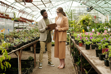 African young landscape designer discussing list of plants together with woman standing in flower...