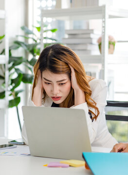 Closeup shot of upset stressed depressed worried unhappy Asian millennial frowning face businesswoman employee sit holding hands on head while working with laptop notebook computer in company office