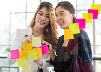 Asian young happy cheerful millennial professional successful businesswoman employee in formal suit standing smiling discussing with colleague writing post it paper on glass board in meeting room