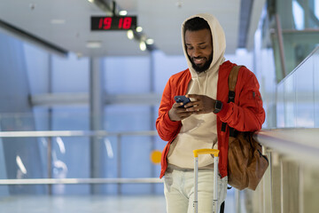Young smiling african american man with luggage in airport chatting in social media before plane boarding, cheerful black guy with smartphone waiting landing on board in terminal, soft focus