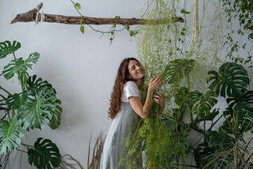 Dreamy curly young woman in grey dress enjoys caring for plants. Plant shop owner hugging plant lovingly. creative millennial woman decorates space with green plants. Floristics, business concept.