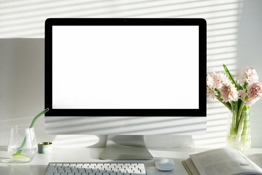 Inspiring workplace for remote work. Desktop computer with blank copy space screen for your promotional content, stationary items, glass of water and fresh summer bouquet. Morning light on the wall