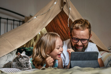 Portrait of a 6 year old boy and his father having fun playing in teepee tent. Father and son using digital tablet watching cartoons or playing computer games lying in kid tent at home. 