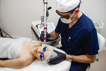young woman in safety glasses getting facial laser treatment by cosmetician in spa clinic. The male...