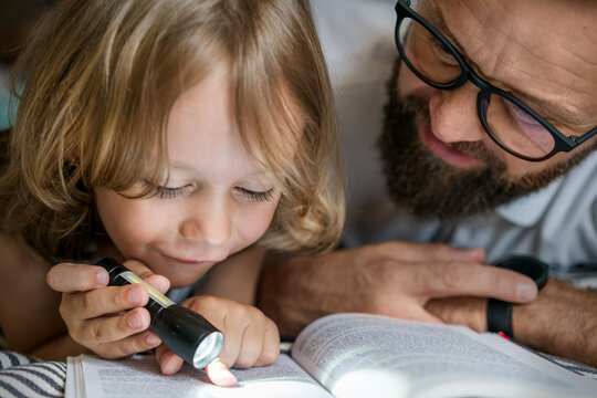 Portrait Of A 6 Year Old Boy And His Father Reading A Book In Teepee Tent. Father And Son With Flashlight Reading Book Under Blanket At Home. 