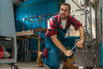 Young male mechanic kneeling on his leg with a smile on his face, while screwing together parts of the metal elements