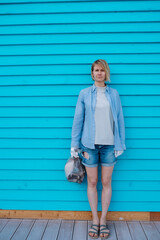 Middle-aged woman with fair hair wearing cotton gloves, standing at blue wooden wall, holding plastic bag with garbage.