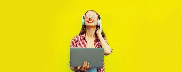 Portrait of modern young woman working with laptop listening to music in headphones on yellow...