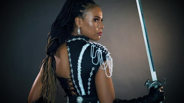 closeup portrait african fantasy woman warrior queen holding sword in hands sharp blade back rear view, sexy face girl princess fashion model posing. black dress creative design, dark gray studio.