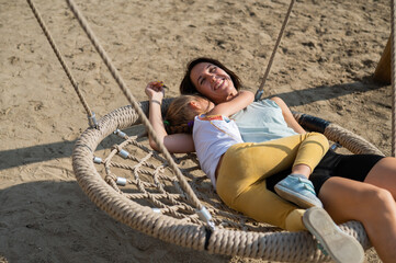 Mom and daughter swing on a round swing. Caucasian woman and little girl have fun on the playground.
