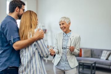 Obraz premium A smiling woman dressed in a suit, holding glasses, shaking hands with a young couple.