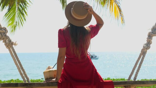 Tourist woman in hat and red dress sitting on wooden swing with young coconut and enjoy sea and palm tree view. Summer vacation in tropical resort. Beachside hotel, yacht on background