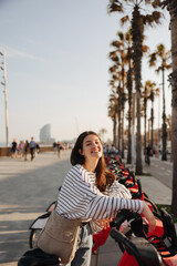 Fototapeta premium Positive young caucasian girl on bicycle parking in city center of Europe. Brunette woman with long hair wears casual clothes. rental concept