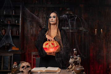 Halloween concept. Black witch holding Halloween pumpkin with carved smily face in hand standing...