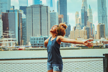 Portrait beautiful young adult woman afro hairstyle with open arms and Manhattan New York City skyline in the background outdoors shot