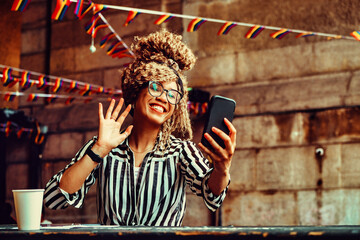 Portrait of smiling woman with afro hairstyle and eyeglasses on a phone video call waving sitting in a coffeehouse shot