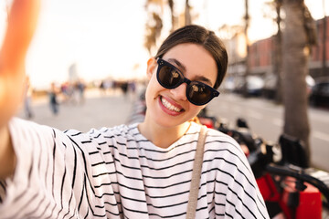 Close-up of young caucasian woman in sunglasses smiling taking selfie in street. Brunette wears striped sweatshirt for walk. People sincere emotions lifestyle concept.