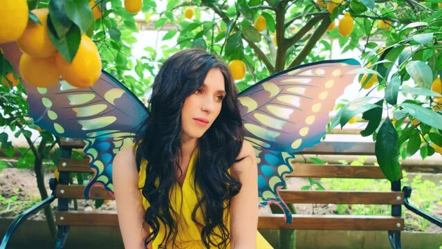 beautiful fantasy fairy woman butterfly resting on wooden bench natural citrus garden, greenhouse. girl sexy face, long flowing hair. Carnival costume, colorful creative moth wings yellow bright dress