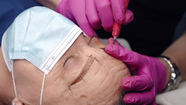 Beautician in sterile pink gloves using tattoo machine for permanent eyebrows correction, female tattoo artist injecting brown colour pigment under the skin of woman for creation eyebrows contour
