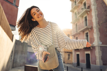 Cheerful young caucasian girl looks at camera, uses wireless technologies on street. Brunette smiles, wears casual clothes and bag. Lifestyle concept
