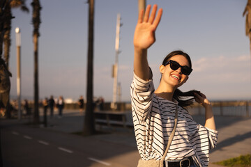 Happy young caucasian woman smiling covering her face with hand standing outdoors in sunny weather. Brunette wears sunglasses, blouse. Relaxation concept