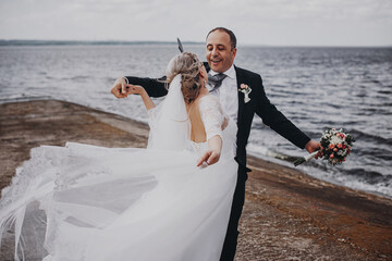 Groom in a black suit with a black bow tie with a beautiful bride in a white long dress embrace and...