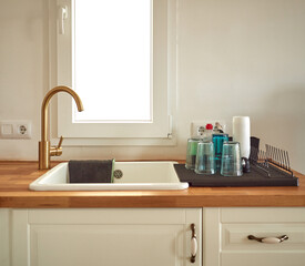 Close up shot of modern white kitchen light furniture sink with rag and golden faucet cozy domestic corner, glasses drying, wooden tabletop, window on background, renovated cooking place design indoor
