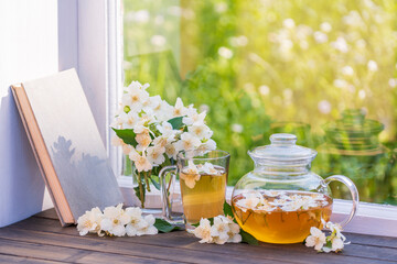 Hot herbal tea in glass teapot, cup and beautiful bouquet of jasmine flowers on windowsill at summer day near garden