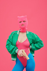 Portrait of fashion model in pink balaclava and body posing with gun against pink background...
