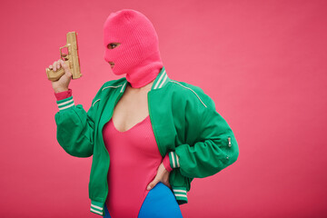 Young criminal girl in pink balaclava and body posing with weapon against pink background - obrazy, fototapety, plakaty