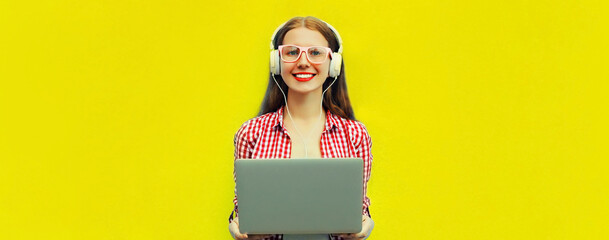 Portrait of modern young woman working with laptop listening to music in headphones on yellow...