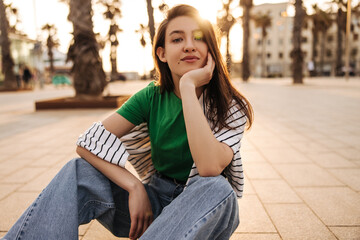 Cute caucasian young woman looking at camera enjoying sunset sitting on pavement in summer. Brunette wears casual clothes. Lifestyle concept