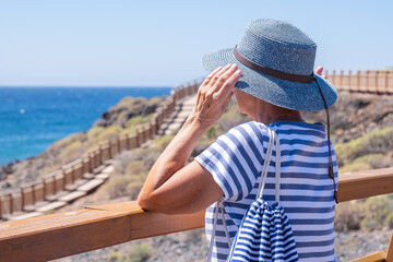 Fototapeta na wymiar Rear view of senior caucasian woman holding her hat while looking at the horizon over water enjoying summer holidays