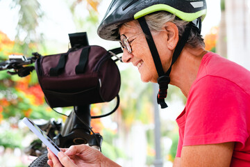 Cheerful cyclist senior woman sitting on a bench in public park close to her bicycle looking at her mobile phone