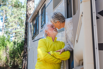 Beautiful caucasian senior couple on the door of their camper van motor home while kissing. Active elderly people enjoying freedom vacation travel in the forest.