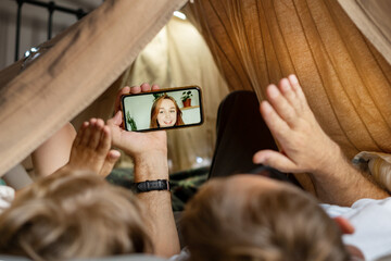 Father and son using smartphone for video call with young woman lying in kid tent at home. Caucasian family in online conversation with each other.