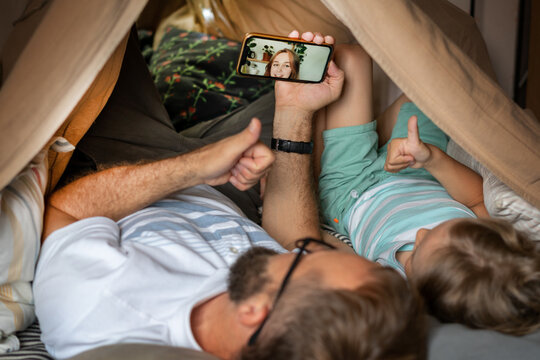 Father and son using smartphone for video call with young woman lying in kid tent at home. Caucasian family in online conversation with each other. - Powered by Adobe