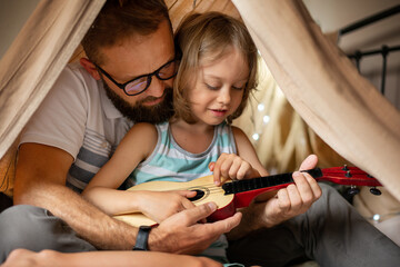 Father and son having fun playing guitar sitting in kid tent at home. Family bonding and enjoying weekend together. Father day.
