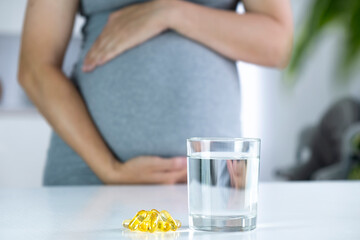 Vitamin D capsules and glass of water and pregnant woman at home interior. Healthy fatty acids...