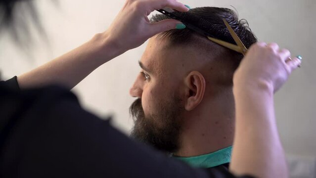 Master cuts hair of men in the barbershop, barber making haircut of attractive bearded man in barbershop using scissors