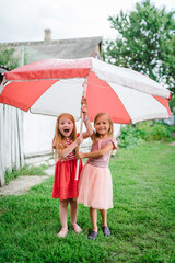 Happy funny childs with big beach umbrella under summer shower or heavy rain in the backyard. Girls walk is wearing dress and enjoying rainfall in spring park. Kids playing and catching rain drops.