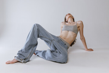 Naklejka premium Portrait of young beautiful girl lying on floor, posing in jeans and cotton underwear top isolated over grey studio background